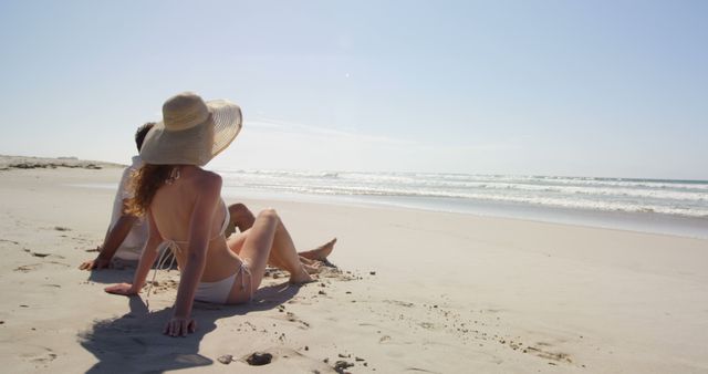 A young Caucasian woman relaxes on a sandy beach, enjoying the sun and the ocean view, with copy space. Her wide-brimmed hat and the serene coastal setting evoke a sense of leisure and summer tranquility.