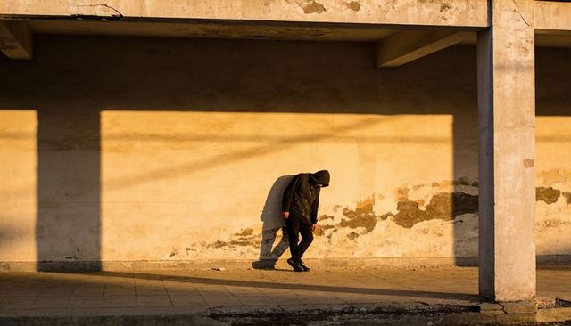 Solitary hooded walker casting long shadows along weathered urban facade at golden hour