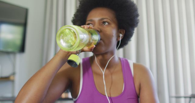 Woman sitting indoors drinking from water bottle after workout session. She is wearing purple activewear and earphones, suggesting an engaged, balanced lifestyle. This image can be used for promotions related to fitness, hydration, healthy living, home workout routines, and active lifestyles.