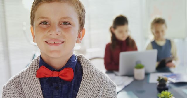 A young Caucasian boy in a bow tie smiles confidently at the camera, with copy space. Behind him, his peers are focused on their work, suggesting a classroom or educational setting.