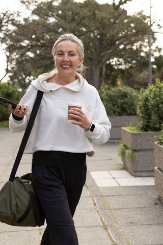 Smiling mature woman walking with coffee and phone, carrying duffel on urban green path