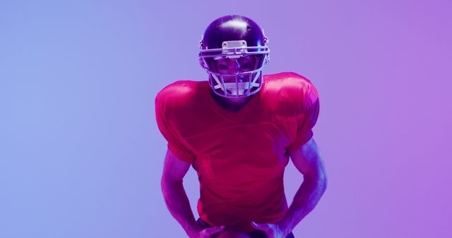 Football player wearing red jersey and helmet posing with colorful lighting can be used for sports advertisements, team posters, fitness blogs, or promoting sporting events. The vivid color emphasizes energy and intensity suitable for conveying action and motivation.