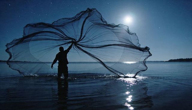 Moonlit fisherman casting circular net over calm water under starry night sky