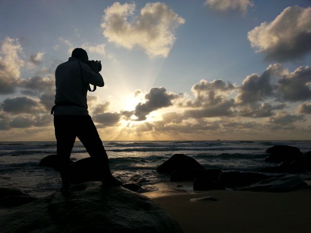 Silhouette of a photographer capturing a stunning sunrise on a rocky beach beside the ocean. Rays of sunlight pierce through clouds, illuminating the scene with a golden hue. Ideal for use in travel promotions, photography websites, and nature blogs.