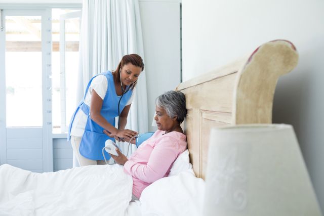 Female doctor measuring blood pressure of senior woman in bedroom. Ideal for use in healthcare, elderly care, home healthcare services, medical checkups, and patient care promotions. Highlights the importance of regular health monitoring and professional medical assistance for seniors.