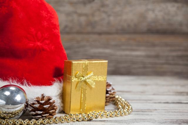 Christmas decorations including a red Santa hat, a gold-wrapped gift, pine cones, a silver bauble, and gold beads are arranged on a wooden table. This festive scene is perfect for holiday greeting cards, seasonal advertisements, and social media posts celebrating the Christmas season.