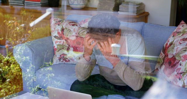 Man sitting on a comfortable sofa with hands on head, showing signs of stress while working remotely on a laptop. Floral pillows behind him create a homey atmosphere. This image is perfect for articles or posts about remote work challenges, mental health, burnout, overwork, work-life balance, and dealing with stress.