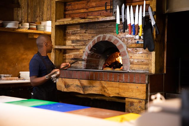 African American male chef working in a restaurant kitchen, using a pizza peel to take pizzas out of a wood-fired oven. Ideal for content related to culinary arts, professional cooking, restaurant operations, and food preparation. Suitable for illustrating articles, blogs, and advertisements focused on gastronomy, chef careers, and restaurant environments.