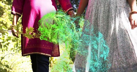 Two indian women walking hand in hand through sunlit garden with teal and green powder
