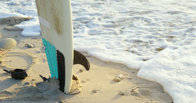 Surfboard stands upright in sand on beach with morning sunlight and ocean waves. Can be used for themes about surf culture, beach vacations, water sports, and morning routines on the coast.