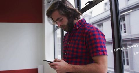 Young man standing by window using smartphone in modern coworking space