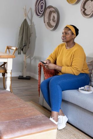 African american woman on sofa holding smartphone in mustard sweater, cozy living room