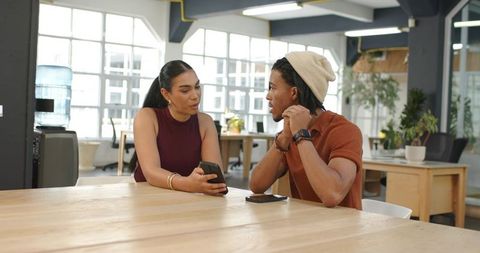Diverse coworkers discussing smartphone at communal table in modern open-plan coworking space
