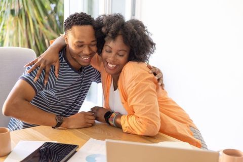 Smiling african american couple hugging while reviewing finances and working on laptop
