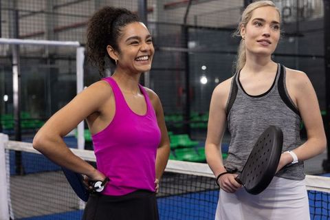Smiling diverse women on padel court holding paddles by net wearing athletic tank tops