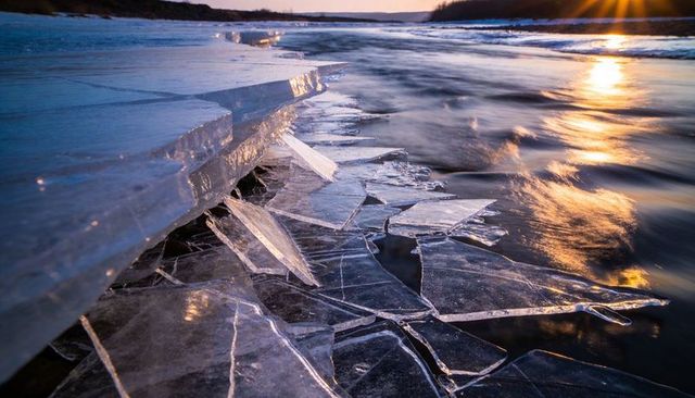 Fracturing ice sheets catching golden sunset over flowing river shattering crystal shards