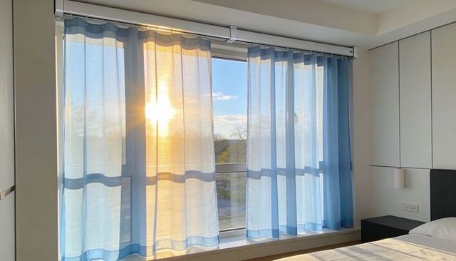 Sunlit bedroom window with sheer blue curtains casting warm morning light across bed