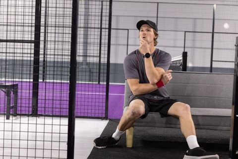 Contemplative athlete sitting on bench in indoor padel court with purple backdrop and mesh fence