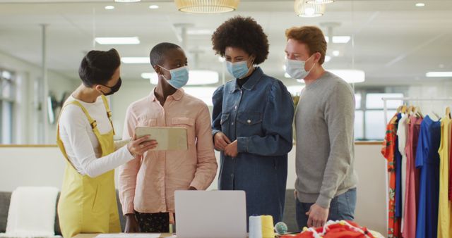 Image shows a diverse team collaborating in a fashion studio while wearing face masks. They appear to be discussing something on a tablet in a modern workspace. The individuals are standing near a desk with a laptop and various clothing items in the background. This image can be used to depict teamwork, creativity, and health precautions in workplaces during the pandemic.