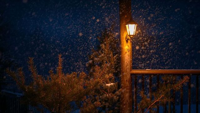 Glowing lantern illuminating snowfall on wooden deck railing with pine branches at night