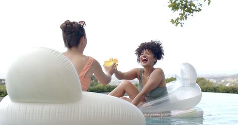Diverse female friends laughing and clinking drinks on pool floats at infinity pool