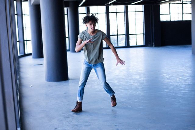 African American transgender man dancing energetically in an empty parking garage. He is wearing casual clothing, including a green t-shirt, jeans, and brown boots. The large windows and concrete pillars create an urban atmosphere. This image can be used to represent themes of gender expression, identity, diversity, and individuality. It is suitable for campaigns promoting inclusivity, freedom, and self-expression.