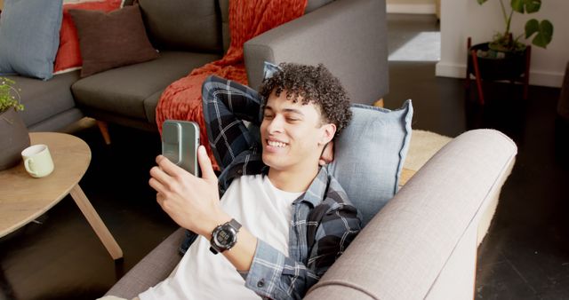 Young man with curly hair lounging comfortably on a couch, smiling while interacting with his smartphone. Modern home interior with cozy furniture in background. Suitable for depicting leisure time, modern communication, casual home environments, and youth lifestyle.