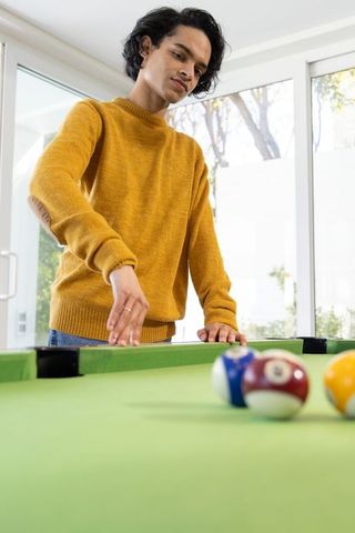 Asian man in mustard sweater reaching for balls on pool table in bright interior