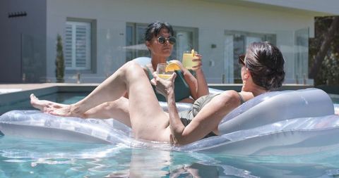 Relaxing women floating on clear loungers sipping citrus cocktails at private pool
