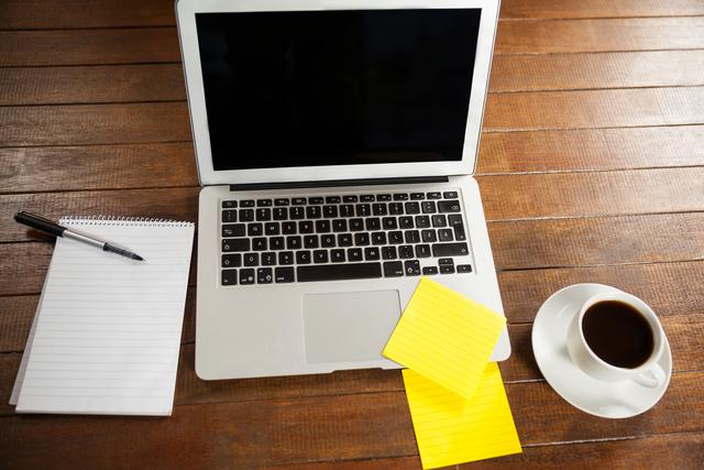 This image shows an office desk with a laptop, notepad, pen, yellow sticky notes, and a cup of coffee on a wooden table. Ideal for illustrating concepts related to productivity, remote work, home office, business, and technology. Perfect for use in articles, blog posts, and presentations about work environments, office setups, and efficient work habits.