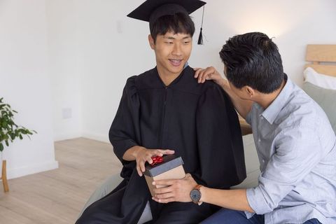 Asian graduate wearing cap and gown receiving gift from father at home