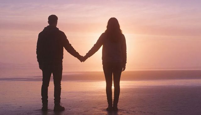 Silhouetted couple holding hands at sunset on calm beach, pastel sky romantic backlit scene
