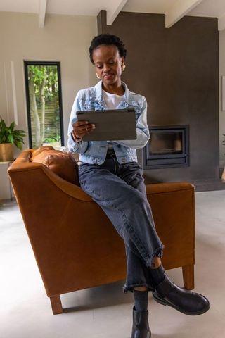 African american woman sitting on leather armchair holding tablet in modern minimalist living room
