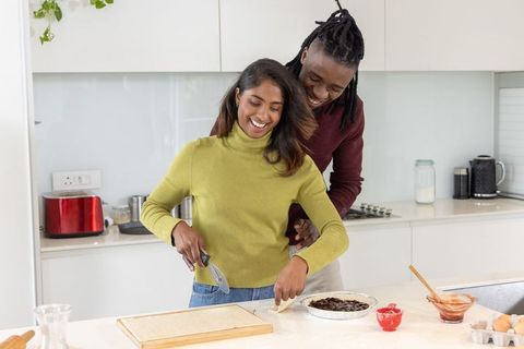 Diverse couple playfully baking at bright modern kitchen island, rolling pastry and filling fruit pi
