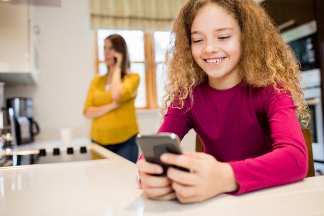Young girl with curly hair smiling while using a mobile phone in a modern kitchen. Woman in the background talking on the phone. Ideal for illustrating family life, technology use among children, and modern home environments.