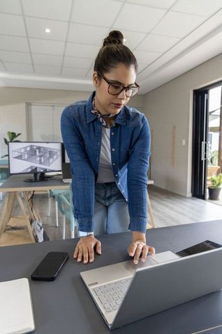 Female architect leaning over desk reviewing 3d building plans on laptop in modern studio