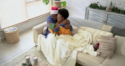 Black couple cuddling under chunky blanket on sofa wearing Santa hat and exchanging gift