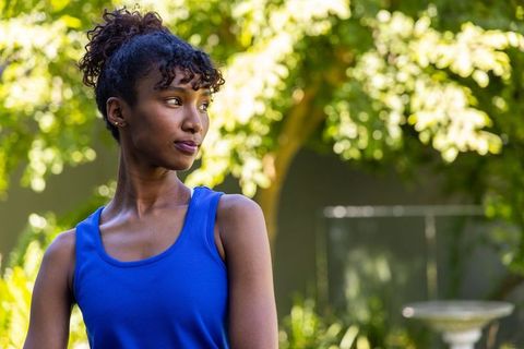 Young woman standing in sunlit garden wearing blue sleeveless top looking thoughtful