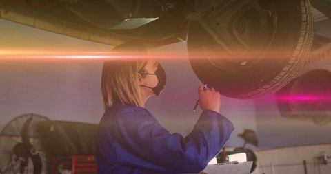 Female mechanic inspecting wheel suspension under raised car wearing mask and coveralls