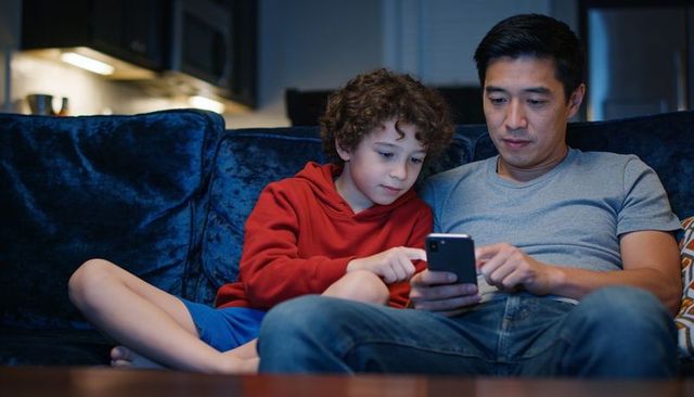 Father and son sharing smartphone on velvet sofa at night, cozy family bonding moment