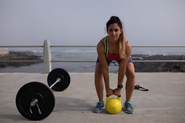 Portrait of a strong Caucasian woman with long dark hair wearing sportswear exercising outdoors by the seaside on a sunny day, strength training lifting kettlebell, looking at camera, barbells and rubber tape next to her.
