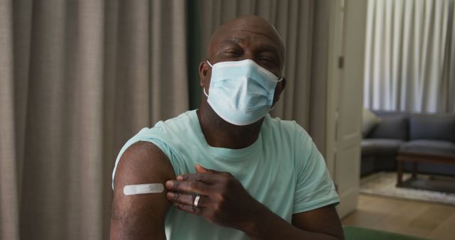 Man sitting on couch, wearing surgical mask, showing vaccination bandage on arm, appearing satisfied and safe. Suitable for use in healthcare promotions, Covid-19 vaccination campaigns, health safety advertisements, and medical care awareness.