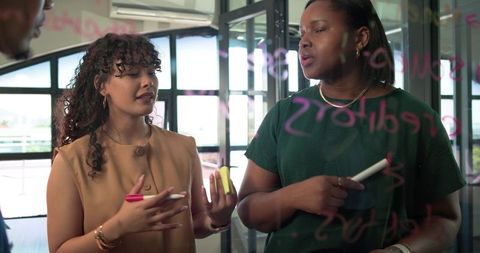Two african american women brainstorming at glass whiteboard in modern office