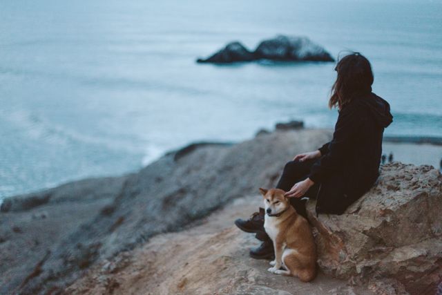 Woman quietly sits on a rocky cliff edge accompanied by a dog, facing the ocean under an overcast sky. This scene evokes a sense of peace and tranquility, making it ideal for use in travel promotions, outdoor adventure content, or advertisements emphasizing serenity and companionship. Perfect for showcasing natural beauty, relaxation, and mindful moments in nature.