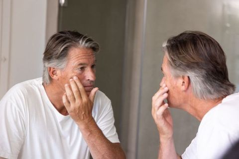 Mature man examining skin in mirror at bathroom vanity, touching cheek, self care