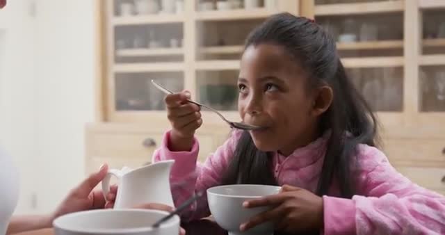 Young girl in pink onesie enjoying cereal at kitchen table while smiling at parent. Portrays warmth and everyday joy of family breakfast moments. Useful for advertisements, parenting magazines, and healthy lifestyle campaigns.