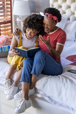 Laughing friends reading journal on bed sharing joyful conversation and relaxed morning