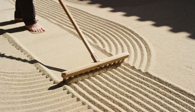 Zen sand raking creating curved parallel grooves with wooden rake, barefoot feet