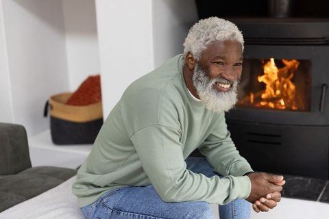 Senior african american man smiling by wood stove, cozy home hearth, warmth and relaxation