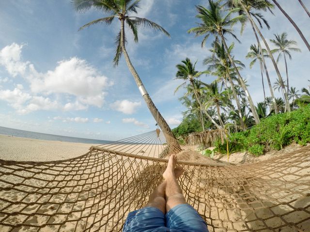 Person relaxing in hammock on tropical beach surrounded by palm trees. Ideal for vacation, travel, and relaxation themes in advertisements, blog posts, and social media content promoting leisure and serenity.